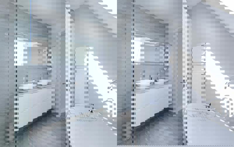 Bilden visar ett modernt badrum i ljusa färger med grå klinkergolv och väggar. Rummet är utrustat med ett stort badkar, dubbla handfat på en vit kommod och en stor spegel. En handdukstork med vita handdukar sitter på väggen, och rummet badar i naturligt ljus från ett takfönster.