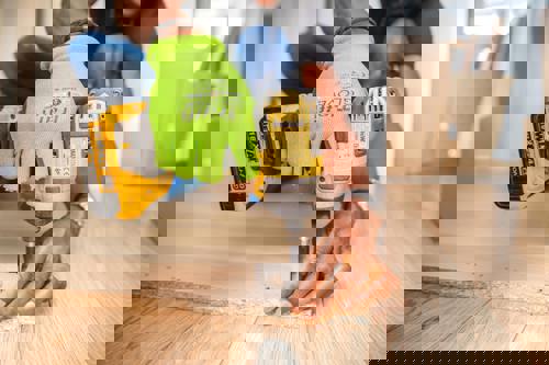 logo Person använder en gul skruvdragare för att montera en metallbeslag på en träskiva, iförd gröna skyddshandskar.