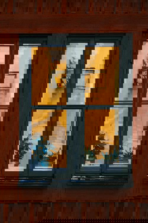 logo Ett fönster i en röd träfasad med mörkgrön karm. Inuti syns krukväxter i fönsterkarmen samt en bokhylla på väggen, upplyst av varmt gult ljus.