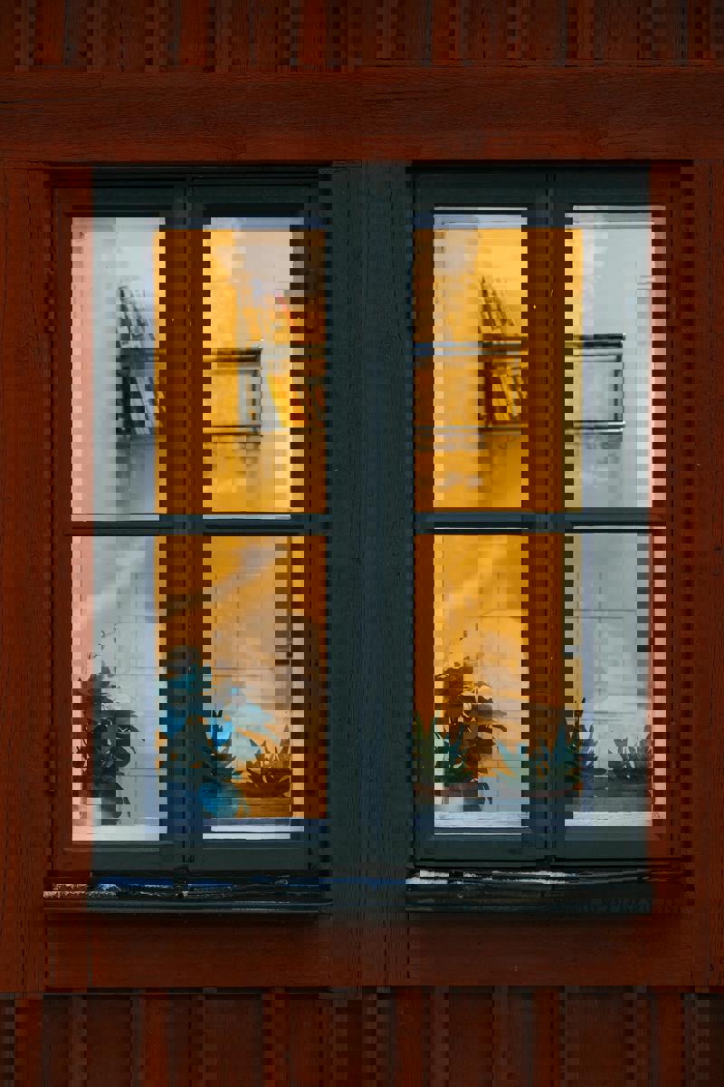 Ett fönster i en röd träfasad med mörkgrön karm. Inuti syns krukväxter i fönsterkarmen samt en bokhylla på väggen, upplyst av varmt gult ljus.