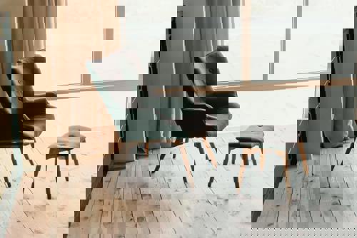 logo En elegant mörkgrå fåtölj med träben och matchande fotpall står på ett ljust trägolv, placerad vid ett stort fönster med utsikt och ljusa gardiner som ramar in rummet.