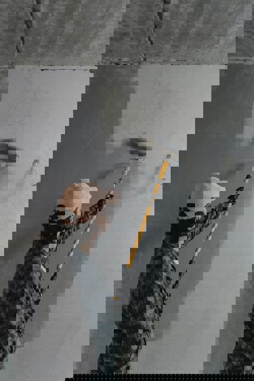 logo En person i rutig skjorta, rosa keps och svarta handskar använder en roller med långt skaft för att måla över en mörk betongvägg i en ljusare färg.