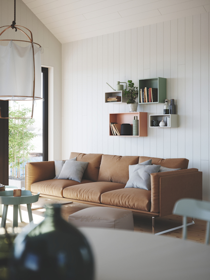 Soffa med kuddar i vardagsrum, hyllor med böcker och växter, ljus interiör med vita väggar och fönster.