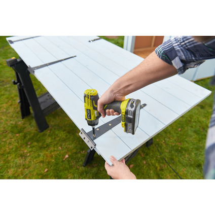 Person använder en grön el-skruvdragare för att fästa en metallgångjärnsskena på en vit dörr som vilar på sågbockar.