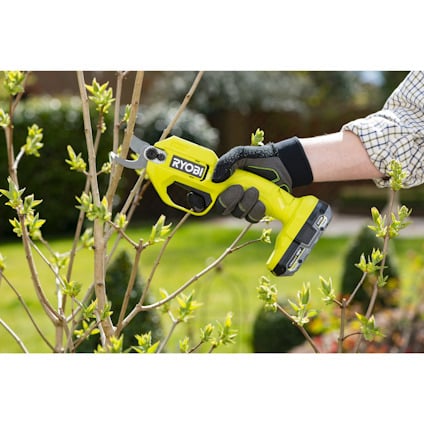 En hand håller en grön RYOBI batteridriven sekatör som klipper en gren.