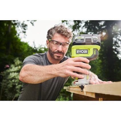 En person använder en grön Ryobi sticksåg för att såga i trä utomhus, bär skyddsglasögon.