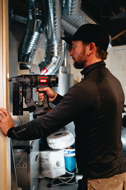 Person i mörk jacka och keps använder en spikpistol för att fästa en träbräda inomhus. Verktyg och ventilation synliga i bakgrunden.