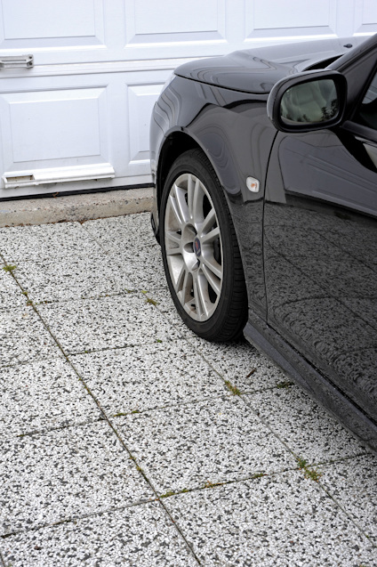 Svart bil parkerad på ljusgrå stenlagd uppfart framför vit garageport.