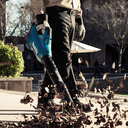 Person använder en blå lövblåsare och blåser bort löv från en trottoar.