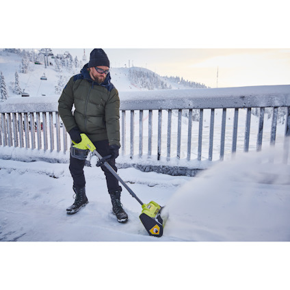 En person med grön jacka och svarta byxor använder en grön snöslunga av märket Ryobi på en snötäckt brygga.