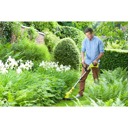 Man klipper gräset med en grön trimmer av märket Ryobi i en lummig trädgård.