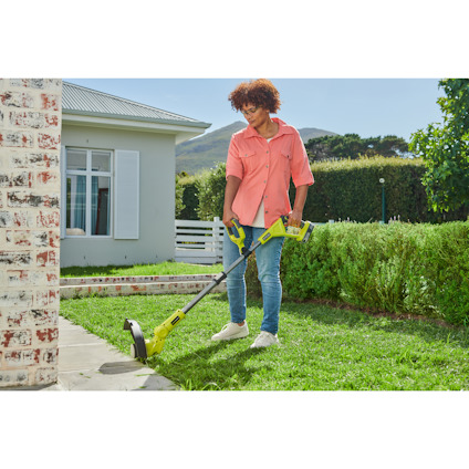 Person använder en grön grästrimmer av märket Ryobi i en trädgård bredvid ett hus.