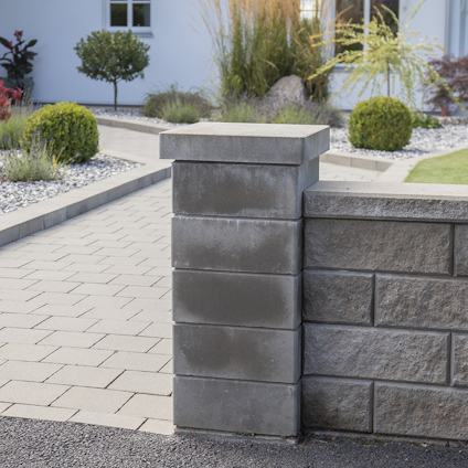 Stenmur vid uppfart med stenplattor och trädgård i bakgrunden.