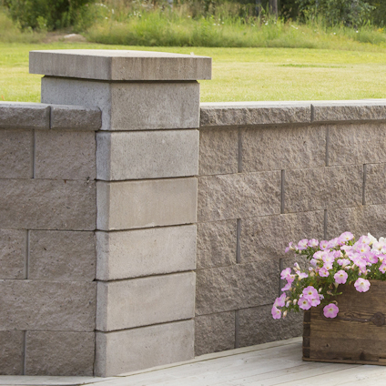 Grå stenmur med dekorativa pelare, omringad av välklippt gräsmatta. En blomlåda med rosa blommor står i förgrunden.