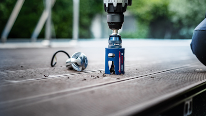 Borrmaskin med blått borrhuvud borrar hål i träplanka, metallspån synliga bredvid.