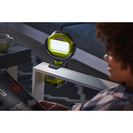 En person håller ett magasin framför en grön RYOBI arbetslampa på ett vitt bord, med en kopp på bordet i bakgrunden.