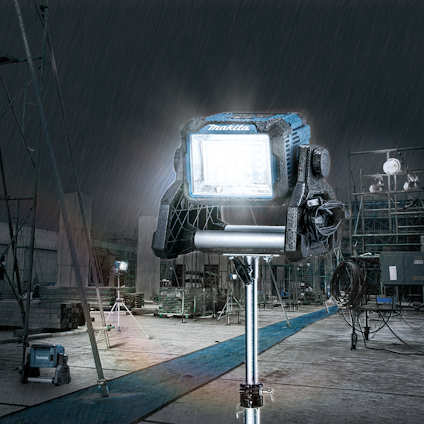 Strålkastare på stativ i regnigt byggområde, omgivna av byggnadsställningar och maskiner.