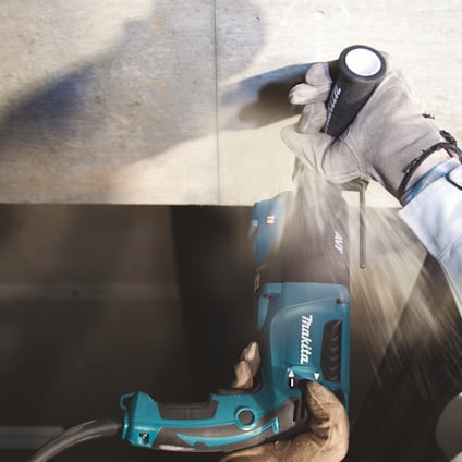 En person i skyddshandskar använder en blå Makita kombihammare vid arbete på en vägg.