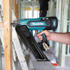 Hand som håller en blå Makita spikpistol vid träkonstruktion i byggarbetsplats.