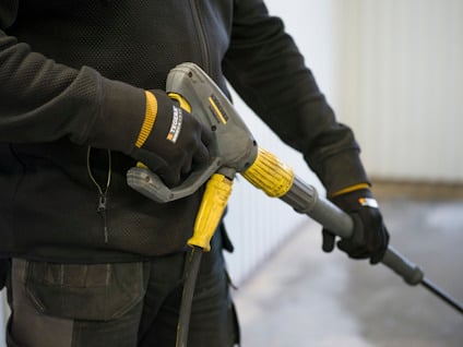 Person håller i en högtryckstvätt med gul och svart handtag, bär handskar och mörka kläder.