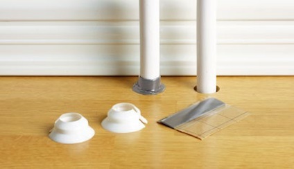 Två vita rör genom ett ljust trägolv, omgivna av vita tätningsringar och en bit aluminiumtejp.