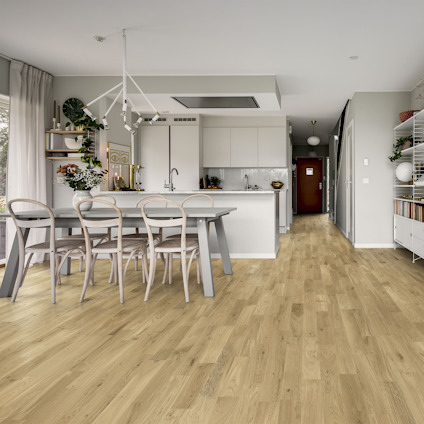 Ljust vardagsrum med ekparkett, matta lack, matbord och stolar. I bakgrunden ett kök med vita skåp och hyllor.