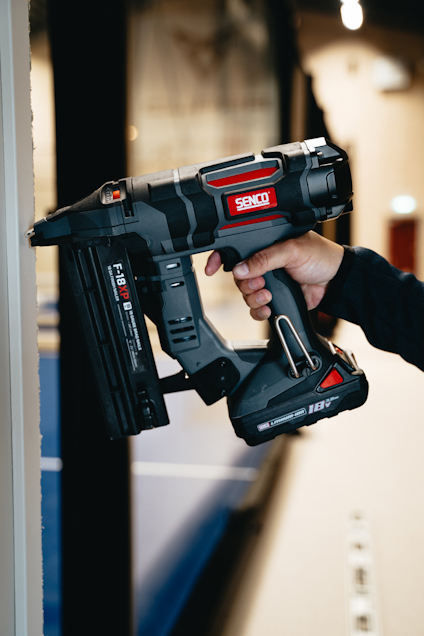 En person håller i en svart och grå spikpistol med märket Senco nära en vägg.