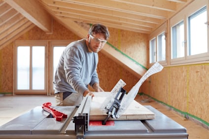 Man using table saw to cut wood planks in an attic under construction. Wearing safety glasses and casual clothing.