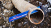 En rostig, cylindrisk metallrör kapas mitt i med ett blått sågblad märkt "Bosch" mot en grusig bakgrund.