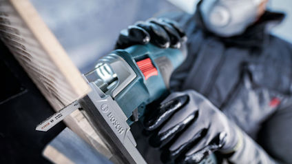 Person i skyddsutrustning använder en elektrisk såg för att såga trä. Bosch-logotyp på sågen.