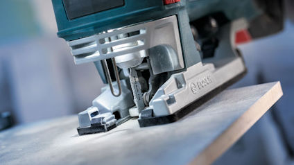 Sticksåg som skär träskiva på ett bord med suddig bakgrund. Märke och modell är otydliga.