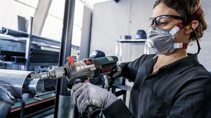 Person i skyddsglasögon och mask använder elverktyg för att såga eller slipa rör i industriell miljö.