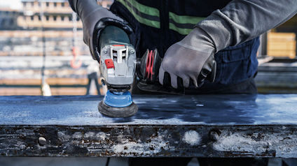 En person slipar en metallplåt med en vinkelslip, iklädd skyddshandskar och arbetskläder.