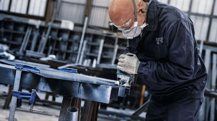 Person slipar metallskiva i verkstad, bär skyddsutrustning och öronskydd.