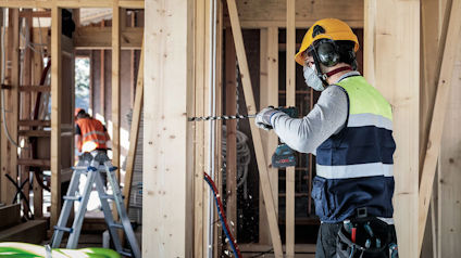 Byggnadsarbetare borrar i träram, iförd skyddsutrustning. En annan arbetare på stege i bakgrunden.