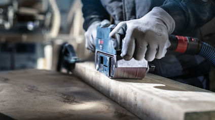 En person slipar trä med ett eldrivet bandslipverktyg, bärande skyddshandskar.