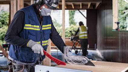 Arbetare i skyddsutrustning skär trä med såg; arbetsplats i bakgrunden.