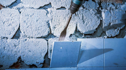 Närbild på en tegelvägg med vit puts, borrmaskin med mejsel används för att ta bort vita kakelplattor.