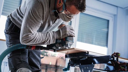 Person med skyddsglasögon och munskydd arbetar med en elhyvel på trä inomhus.