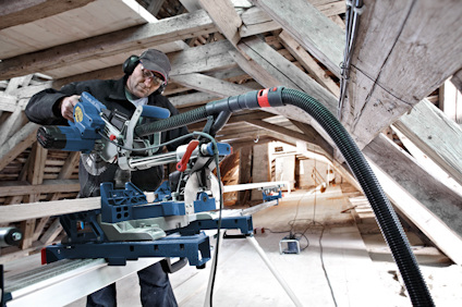 Person med hörselskydd använder elektrisk kap- och gersåg i byggnad med träbjälkar.