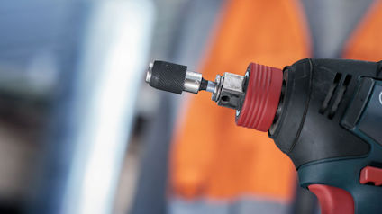 Borrmaskin med fastspänd borrchuck, suddiga orange arbetskläder i bakgrunden.