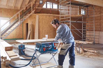 Man arbetar med en cirkelsåg i ett trähus under uppbyggnad, omgiven av byggmaterial och ställningar.