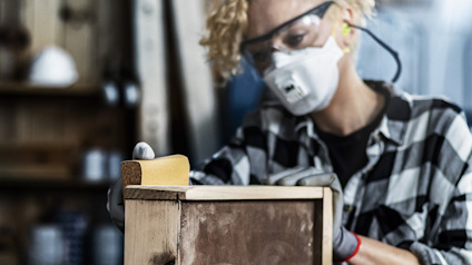 Person i skyddsmask och glasögon slipar en träyta med ett sandpapperblock.
