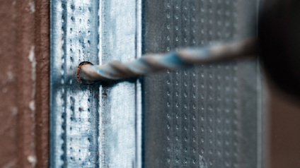 En borr träffar en metallplåt, nära en svetsfog, med en borrspets som just börjat borra.