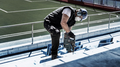Person i arbetskläder borrar i marken på en stadion med konstgräs i bakgrunden.