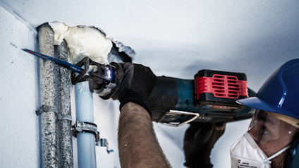 Person i skyddsmask och hjälm använder sågliknande verktyg på rör nära tak.