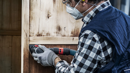 Person med skyddsmask och verktyg slipar en trädörr.
