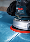 Två händer håller en Bosch GEX 125-150 AVE Professional excenterslip på en blå yta.